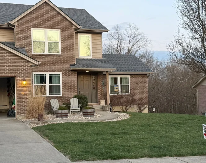 A two-story brick house with a garage and a green lawn. There are two chairs and plants by the front door.